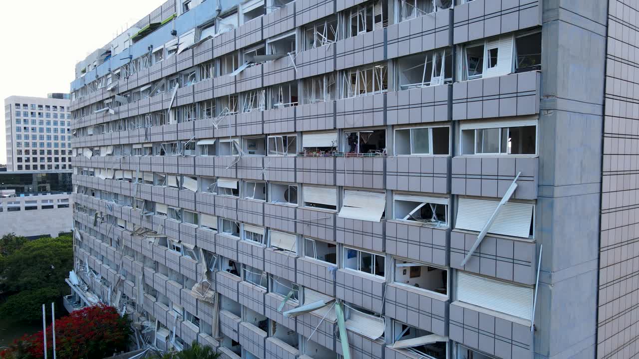 A lateral drone shot moves from left to right, capturing the full façade of a residential building in Tel Aviv severely damaged by an Iranian missile. Windows are shattered, debris is visible