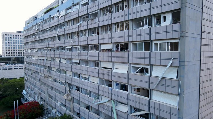 Una ripresa laterale con drone si muove da sinistra a destra, catturando l'intera facciata di un edificio residenziale a Tel Aviv gravemente danneggiato da un missile iraniano. Le finestre sono in frantumi, i detriti sono visibili.