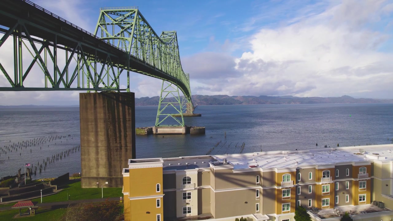 Um drone paira ao lado da Ponte Astoria, oferecendo uma vista panorâmica do Rio Columbia abaixo. Nuvens brancas e fofas flutuam pelo céu, emoldurando a ponte e o rio.