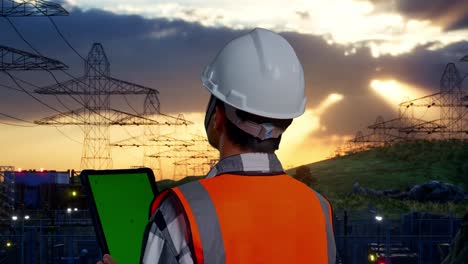 close up back view of asian male engineer with safety helmet working on a green screen tablet and looking around while standing near high voltage tower