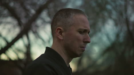 a man in a green jacket smokes a cigarette outdoors at dusk, with smoke visibly escaping his nose and mouth