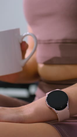 vista parcial de la parte inferior del cuerpo de una mujer en traje de gimnasio usando un reloj inteligente mientras bebe agua, momento relajado después del entrenamiento con fondo borroso con varios equipos de gimnasia
