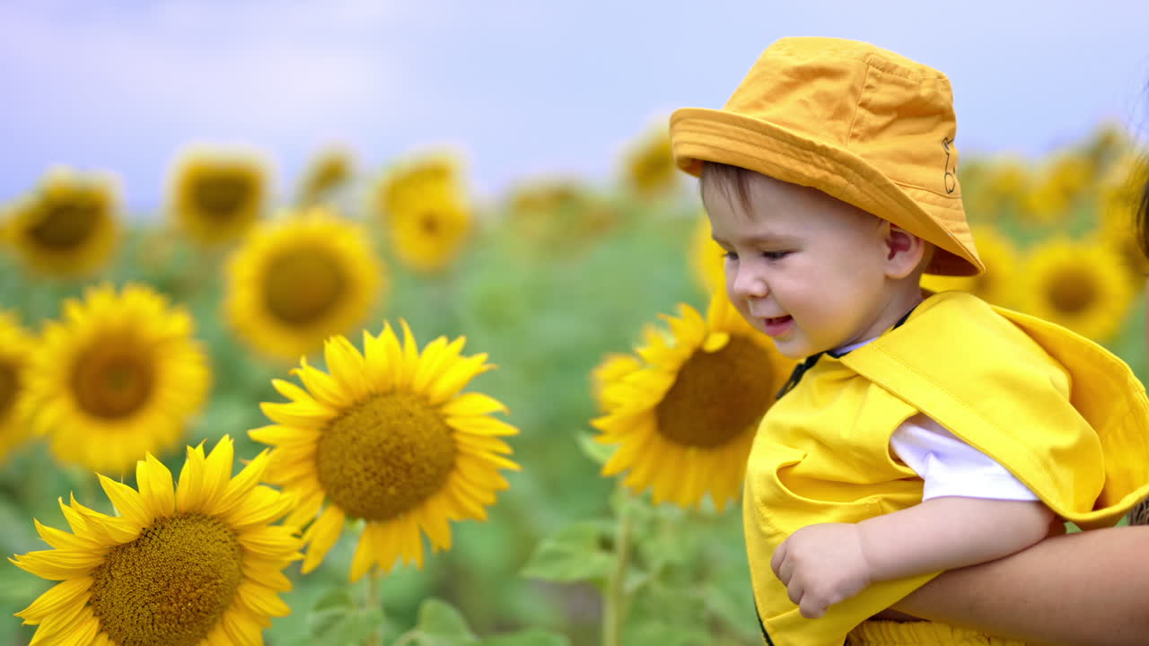 Smiling adorable kid enjoying flowers around him. Mother is holding her son in hands while he touches the sunflowers with interest.