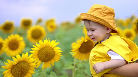 smiling adorable kid enjoying flowers around him. mother is holding her son in hands while he touches the sunflowers with interest.
