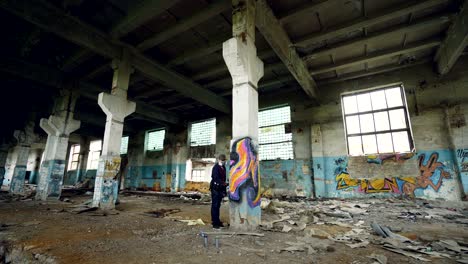 male graffiti artist in respirator is shaking spray paint then painting on high pillar inside dirty empty building. beautiful abstract image on column is visible.