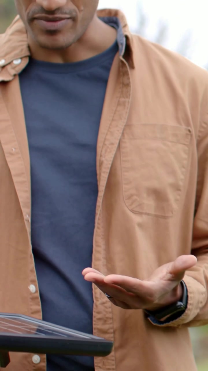 Video verticale: Uomo con camicia color cachi e maglietta blu navy che tiene un cavo di un pannello solare al parco, con la telecamera che si alza