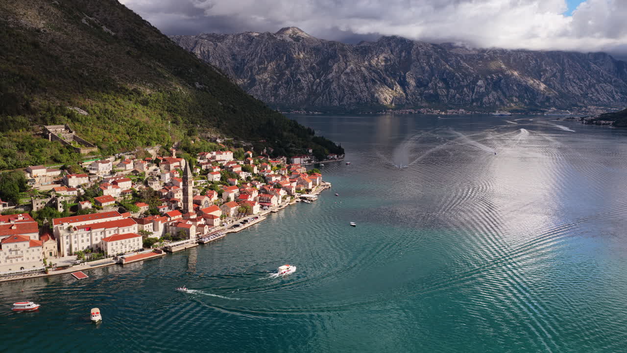 Pemandangan udara dari drone di kota lereng bukit Perast, menyoroti menara gereja yang tinggi, rumah-rumah beratap merah yang padat, dan tepi laut di sepanjang Teluk Kotor