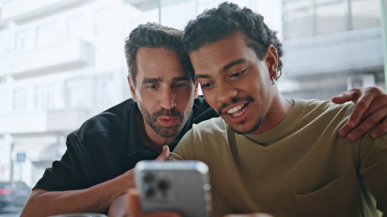 Coppia gay che saluta in videochiamata tramite smartphone in un caffè, primo piano. Incontro online.