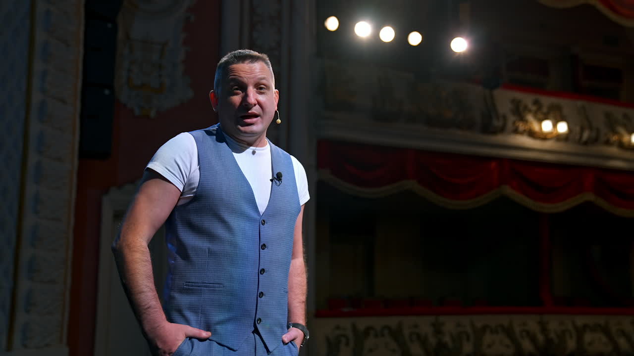 Portrait of an actor during the rehearsal. Man in grey suit standing on stage in the empty theater and talking to camera. Lockdown and world crisis.