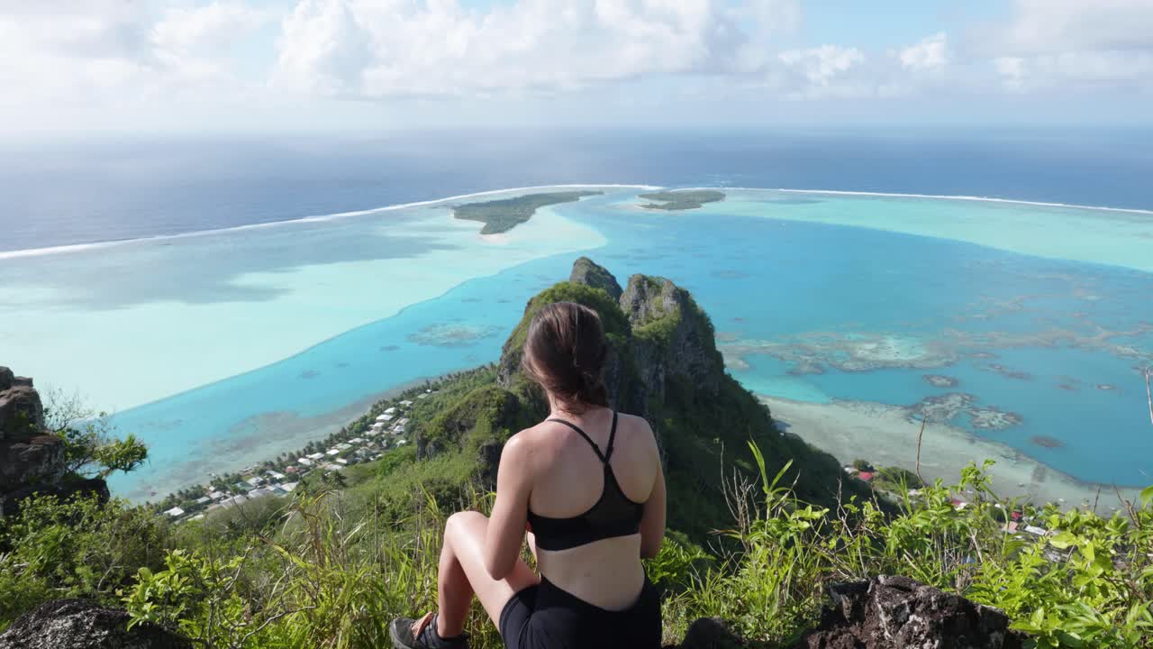 Young caucasian woman in sport clothes and sunglasses sitting at a lagoon and coral reef viewpoint in a green tropical pacific island on a sunny day at Mount Teurafaatiu, Maupiti, French Polynesia.