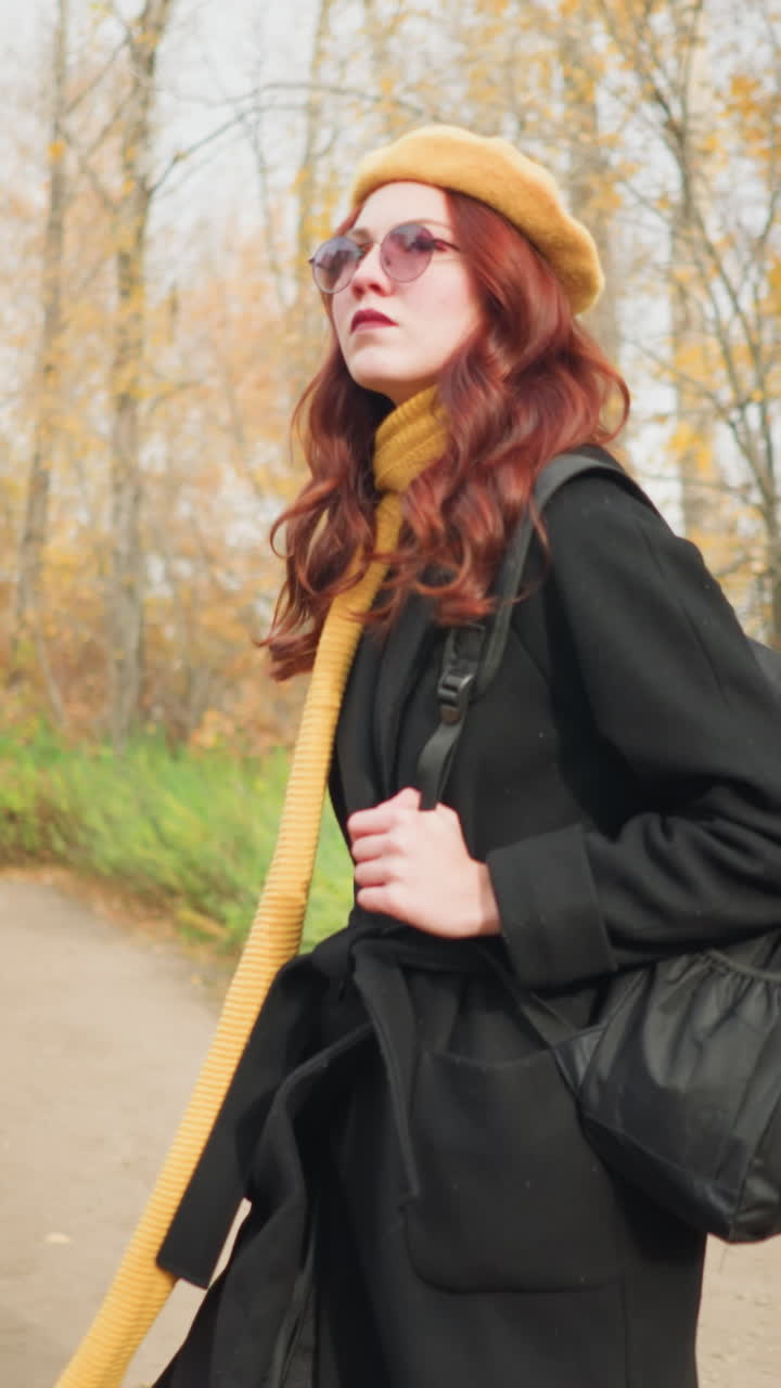 검은 코트, 노란 스카프, 베레트를 입은 관광객이 조용한 가을 공원에서 목조 게지보에서 나와, 집중된 표정으로 가방 줄을 들고, 배경에 목조 벤치를 들고 있습니다.