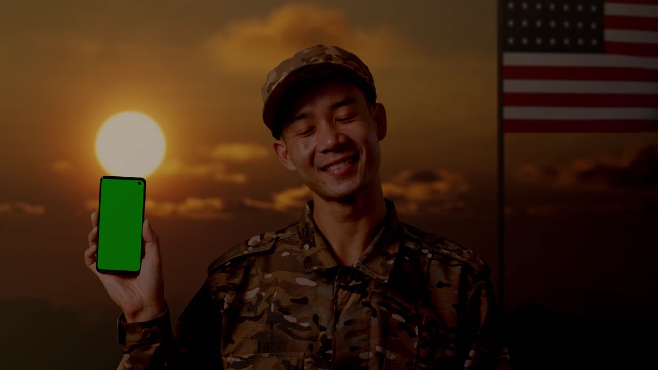 primer plano de un soldado asiático sonriendo y mostrando un teléfono inteligente de pantalla verde a la cámara mientras está de pie con la bandera de los estados unidos, hora del atardecer
