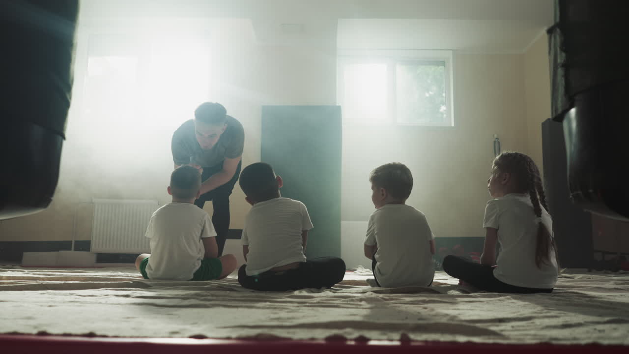 훈련사는 무술 클럽에서 나쁜 학생을 비난합니다. 어린 아이들은 체육관에서 바닥에 앉아있는 전문 강사를 듣습니다. 느린 동작. 전투 수업