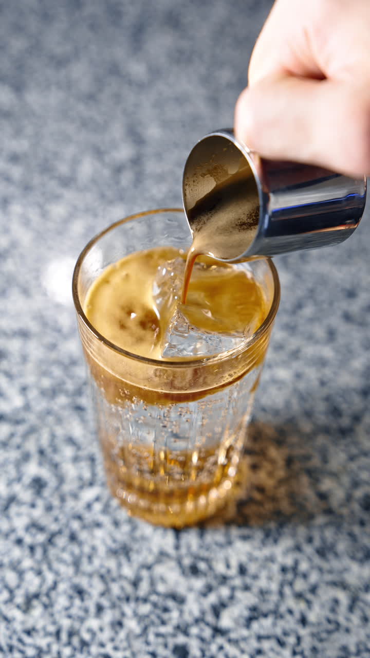 A glass filled with fizzy beverage and ice cube. Male hand pours some fresh coffee from a metal measure cup. Close up. Vertical video.