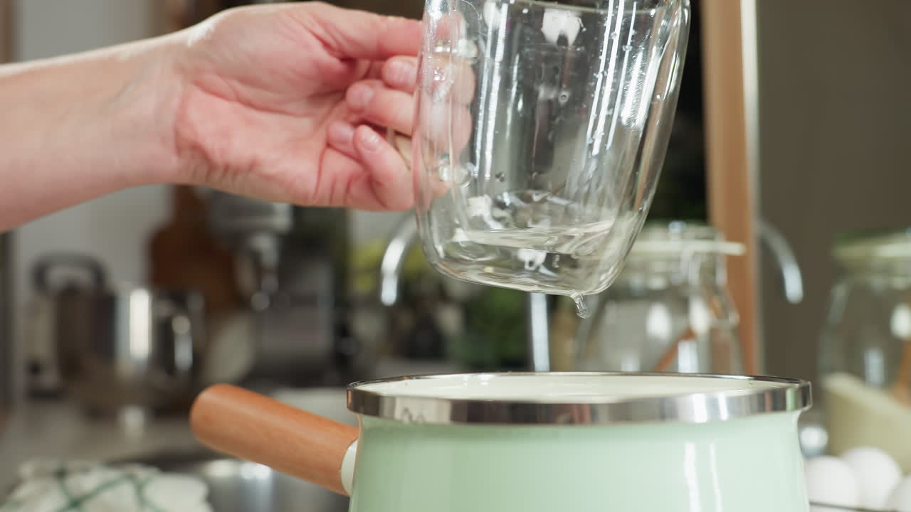 Clean water being poured from clear glass into green jar with wooden handle while chef prepares to drop cup back on kitchen counter, surrounded by utensils, jars and eggs