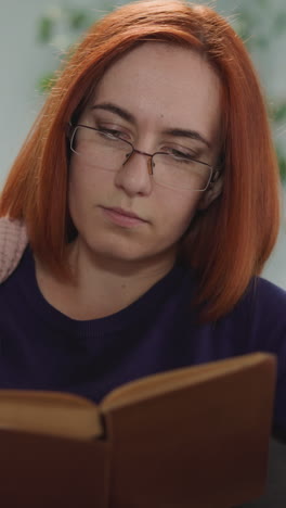 concentrated housewife in glasses reads book flipping pages. woman with ginger hair focuses on captivating story sitting in living room at home closeup