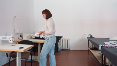 fashion designer in jeans and plain long sleeve top approaches sewing machine holding white fabric from large table, talks to stitching machine while placing fabric on it ready for sewing process