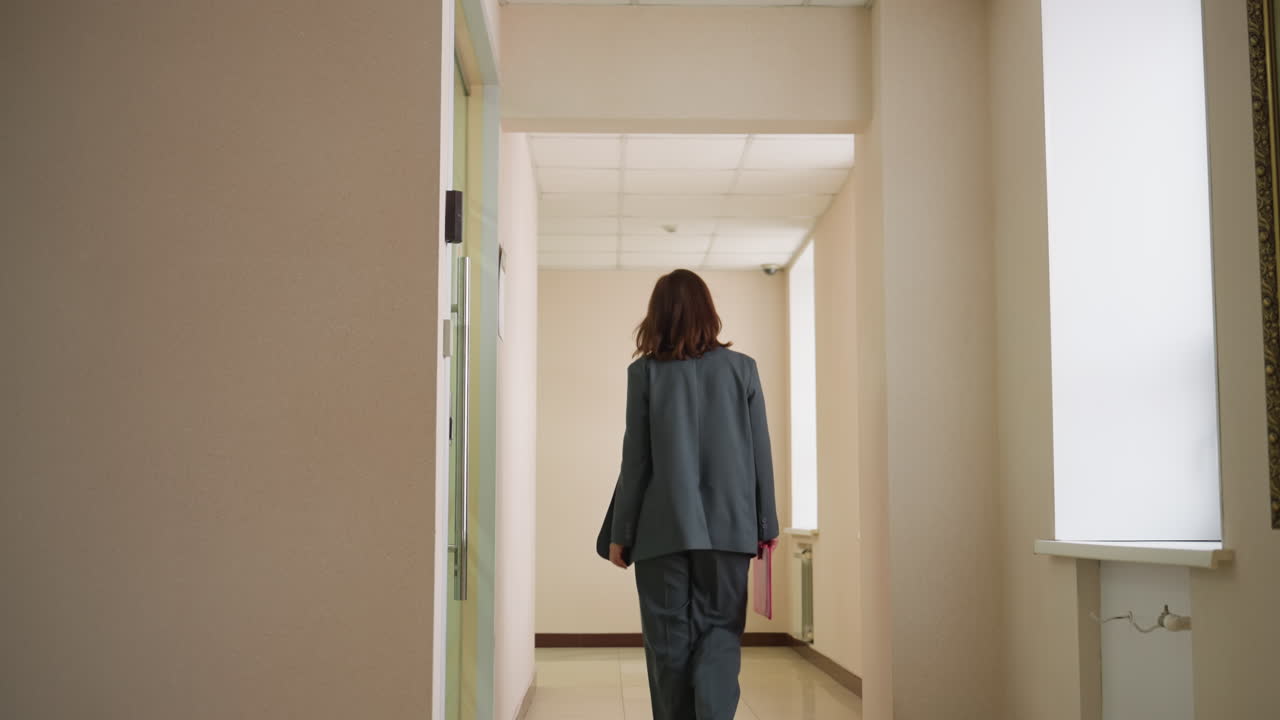 Rear view of confident businesswoman walking alone down quiet office corridor in formal suit toward meeting room, suggesting ambition, purpose, career progression, professional mindset