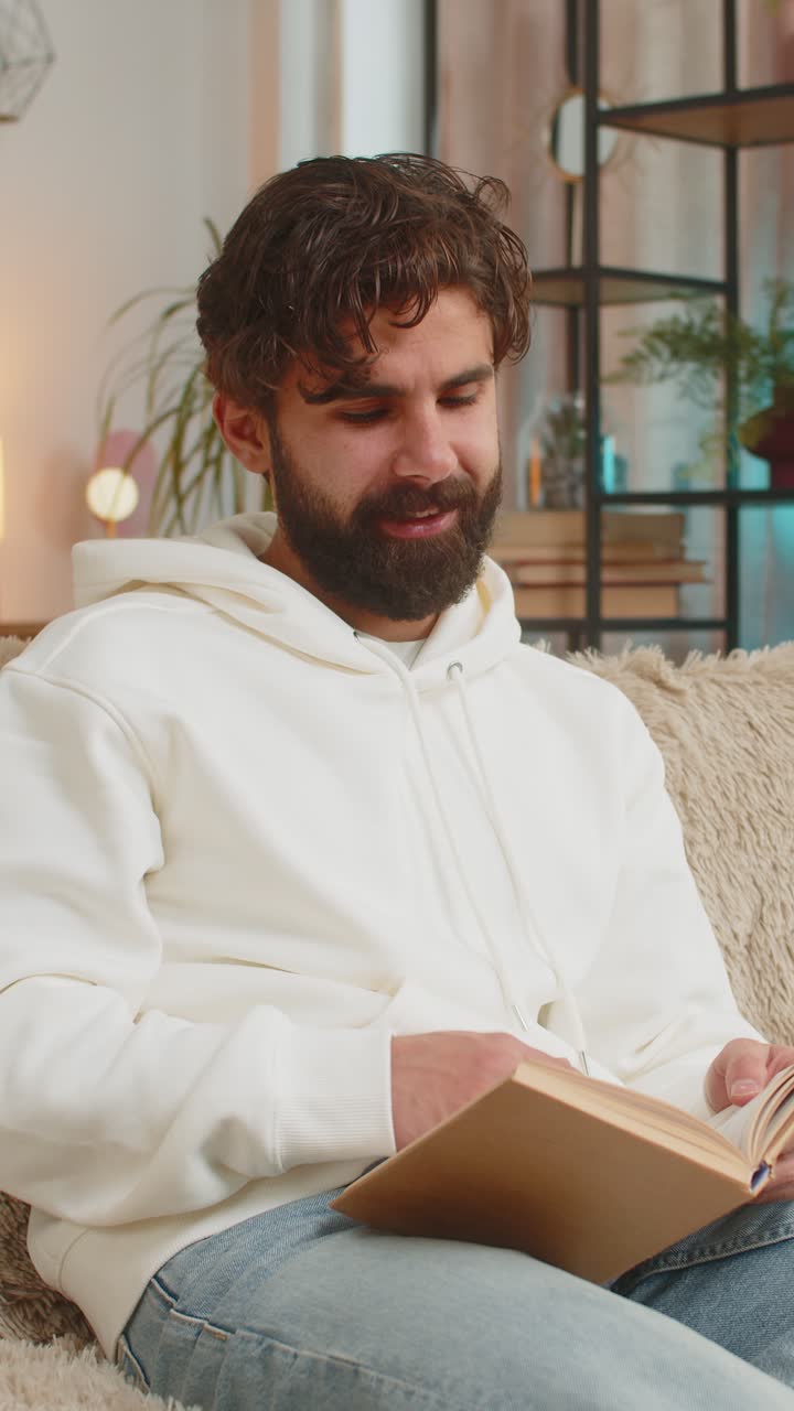 Indian man reading interesting book turning pages smiling enjoying literature sitting on sofa