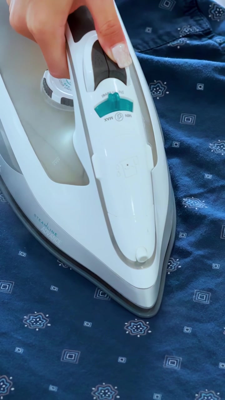A person uses a steam iron to smooth wrinkles on a blue patterned shirt in bright, natural lighting. Close-up, steady camera, household setting