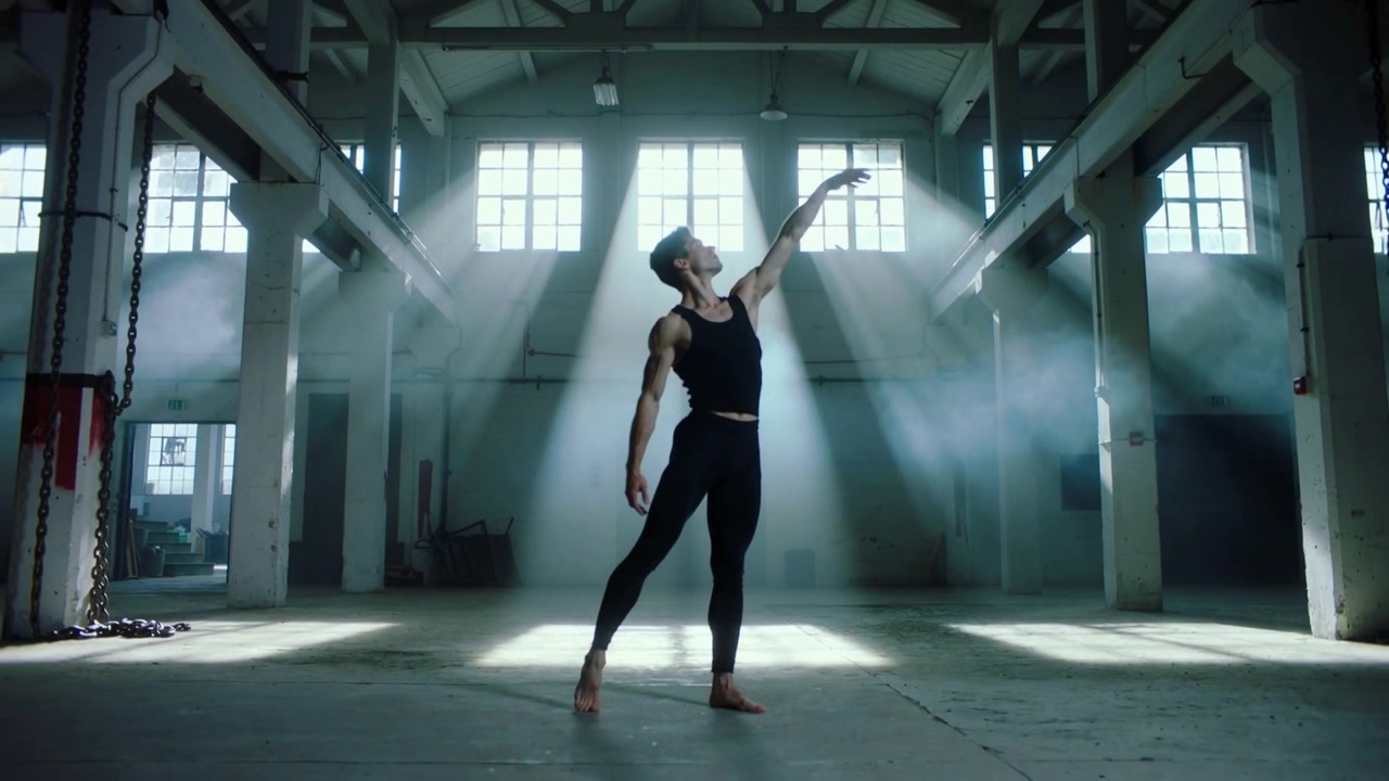Starting dancer in black outfit shifting weight, reaching up, forming line in loft with light beams