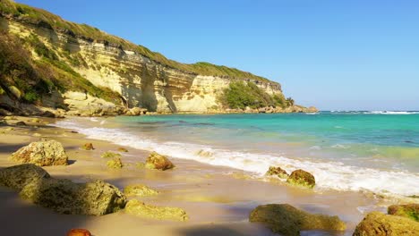 breathtaking low flight above sandy rocky beach towards turquoise ocean waves and limestone sheer cliff mountain in background on cloudless blue sky day, overhead aerial approach