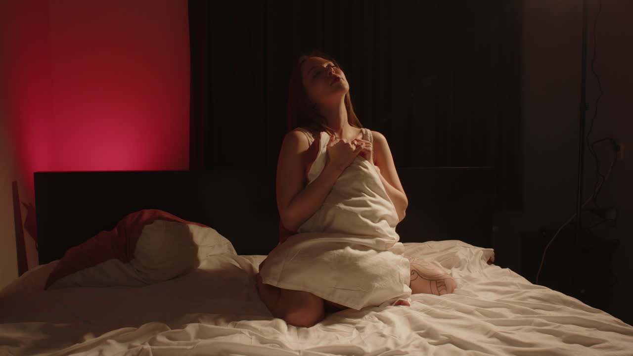 Cinematic wide view of a beautiful woman sitting on her knees in bed wearing white lingerie and holding a pillow in front of her attractive body