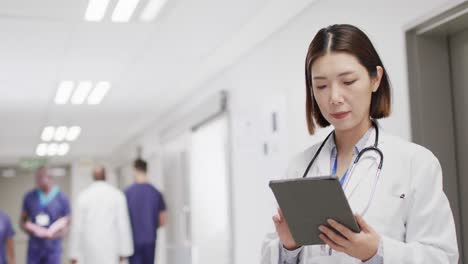 video portrait of smiling asian female doctor with tablet in hospital corridor, with copy space