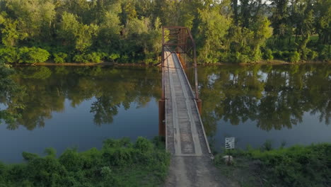 Toma-Aérea-De-Izquierda-A-Derecha-De-Un-Viejo-Puente-De-Hierro-Sobre-Un-Río-Rural-Bordeado-De-árboles-En-La-Cálida-Luz-De-La-Tarde