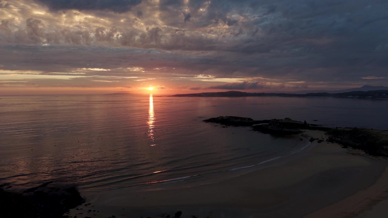 Dronekuva hehkuvasta auringonlaskusta tyynen meren yllä, auringon heijastuessa Paxoksen rannalla ja sen kallioisella niemellä Galiciassa, Espanjassa