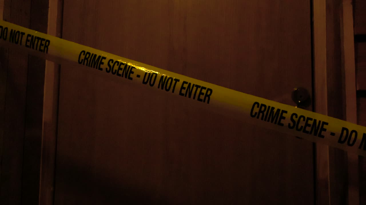 Police investigation forensic yellow Crime Scene Do Not Enter tape ribbon pulled in front of a wooden door at an apartment or a house while lights are dim and the tape moves slightly