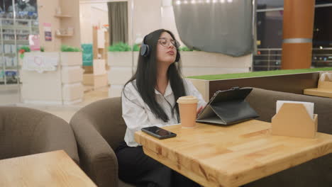 young woman with headphones and glasses seated in modern cafe attentively working on tablet with coffee on table, focused on task with calm expression and cozy professional atmosphere around her