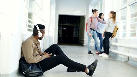 joven estudiante afroamericano guapo sentado en el suelo en el corredor blanco con auriculares en la cabeza escuchando música sosteniendo un teléfono inteligente enviando mensajes de texto a alguien. sus compañeros de clase están detrás de él