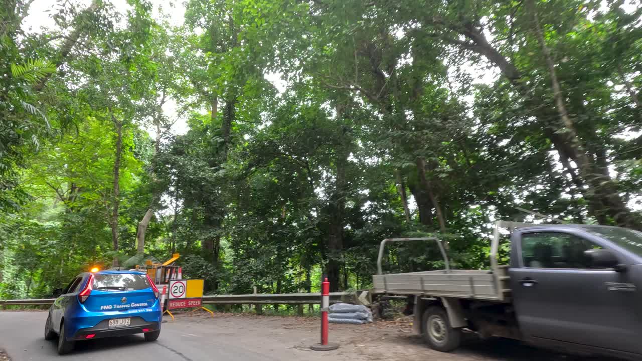 Blue car drives past roadwork vehicles on narrow rainforest road, natural daylight, steady camera