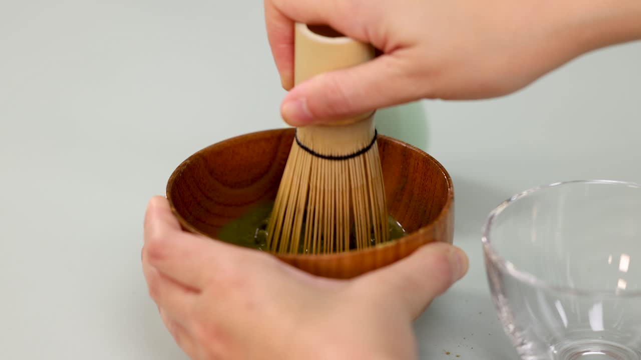 Hands whisk matcha green tea in wooden bowl using traditional bamboo whisk, bright soft lighting