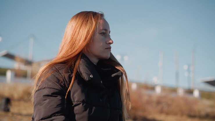 Donna con capelli lunghi sta in piedi all'aperto in un ambiente naturale, indossando un piumino nero. La scena presenta una luce diurna soffusa e uno sfondo paesaggistico aperto con strutture distanti visibili sotto un cielo sereno