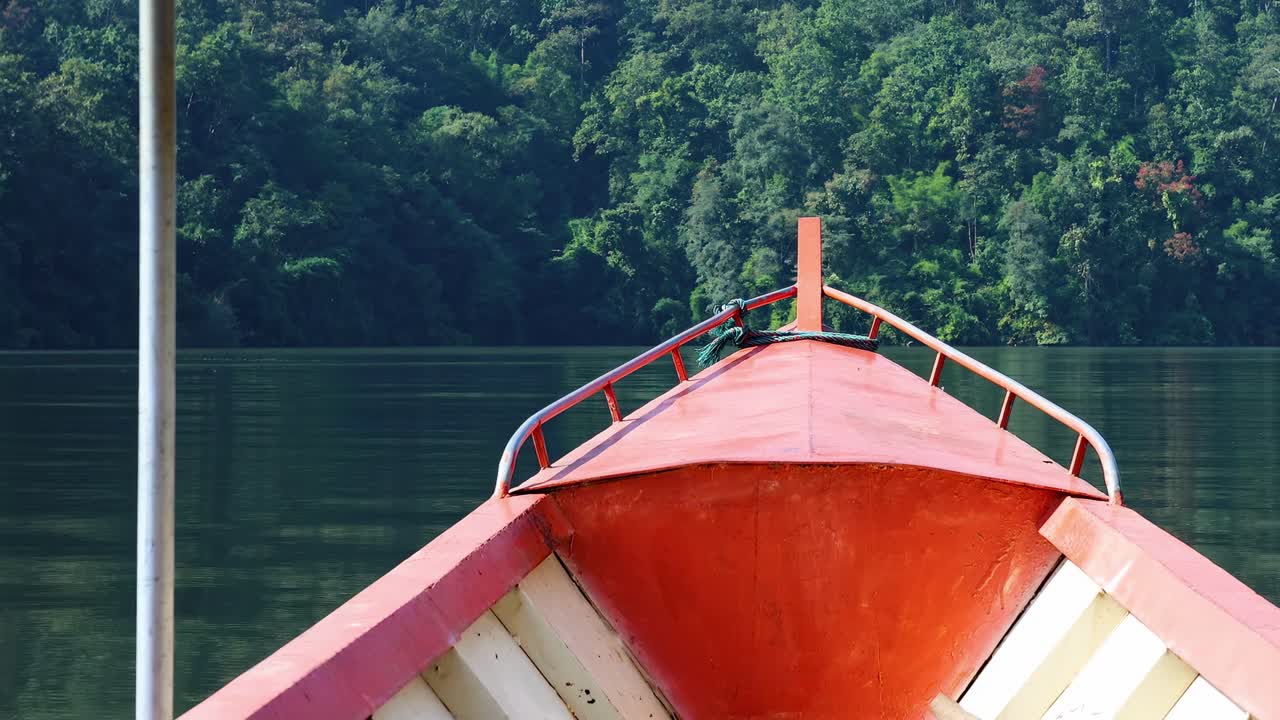 A red boat's bow glides smoothly across a serene lake with lush green trees in the background.