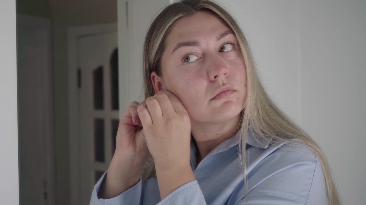 Job seeker in a pale shirt carefully fastens stud earrings, completing her grooming routine for a remote video meeting.