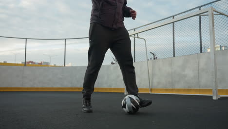 man practicing soccer drills on sport arena, demonstrating ball control and focus under soft daylight, background includes urban residential buildings, lampposts, mesh fencing, and a goalpost