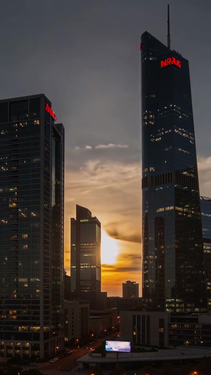 Video verticale: Sole che tramonta tra le torri, torre di vetro che illumina un logo rosso e le finestre