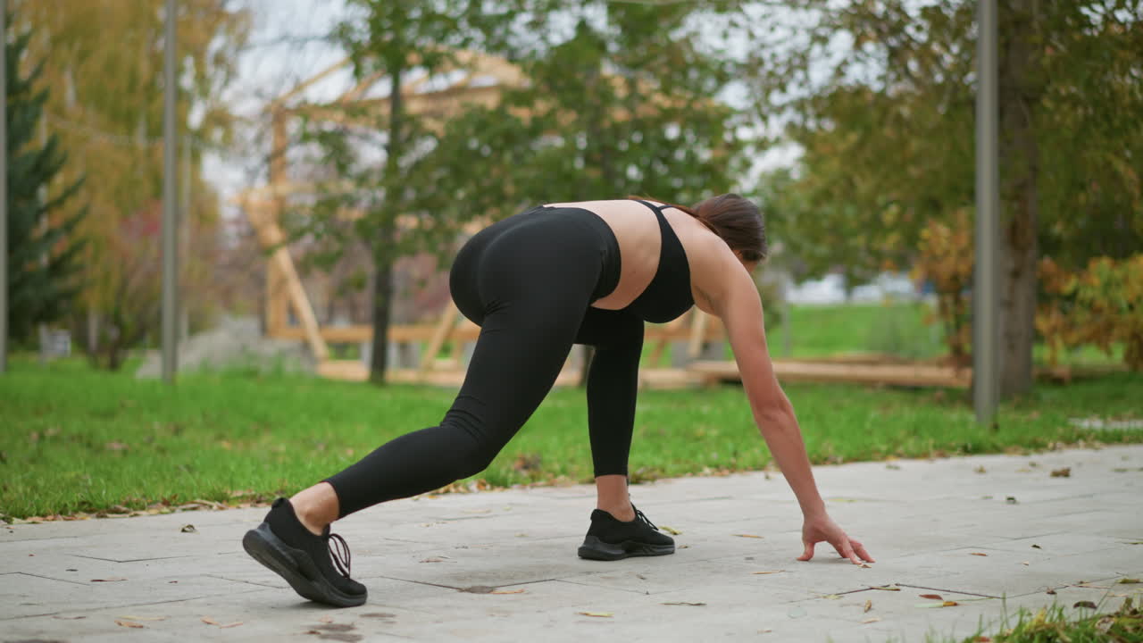 Sport woman in black leggings training outdoors in park, shes preparing to run with hands on the ground, focusing on her form, with trees and iron fence in background
