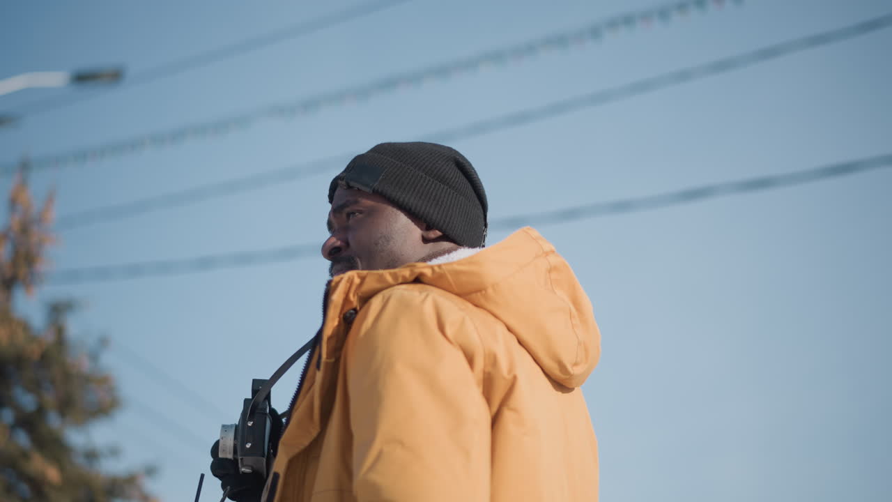 back view reporter holding beverage while turning to shoot with camera under bright winter sun on blur urban street wearing gloves beanie jacket near power wires on snow covered ground capturing