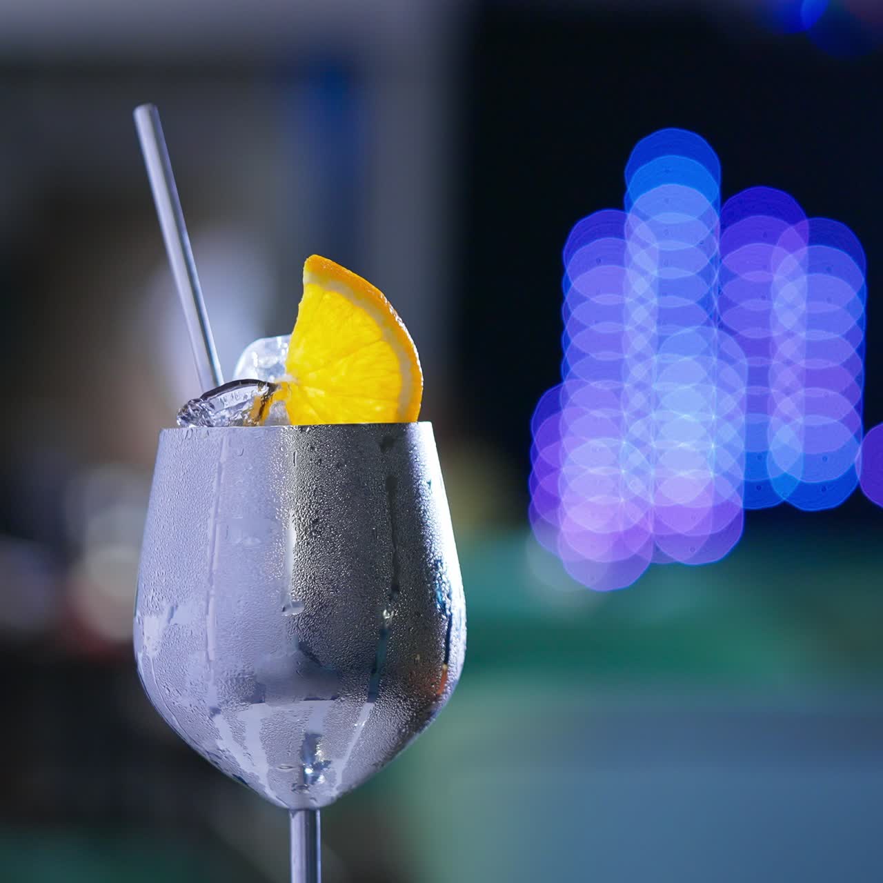 Rotating metal wineglass with misted surface. Cocktail with ice and orange slice on top and metal straw inside. Close up. Blurred backdrop