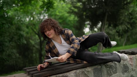 confident brunette girl with curly hair lies down on a bench while writing down her ideas and preparing for exams in the park