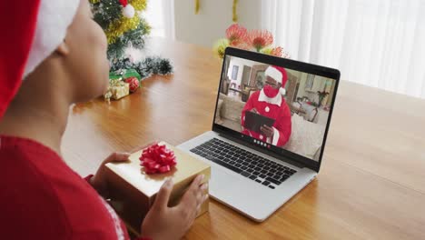 Mujer-Afroamericana-Con-Gorro-De-Papá-Noel-Usando-Una-Computadora-Portátil-Para-Una-Videollamada-Navideña,-Con-Papá-Noel-En-La-Pantalla