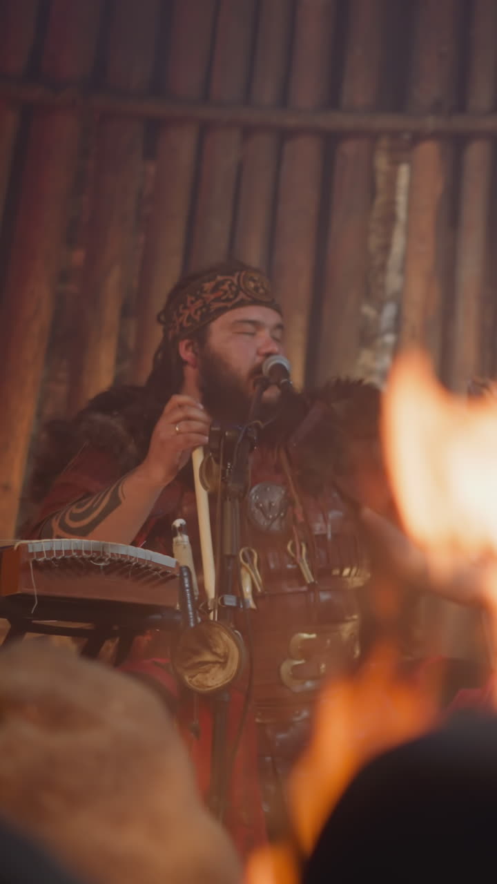 Plump man sings while friend plays drums in traditional Altai house with fireplace. Rock band performance at atmospheric concert. Traditions and life