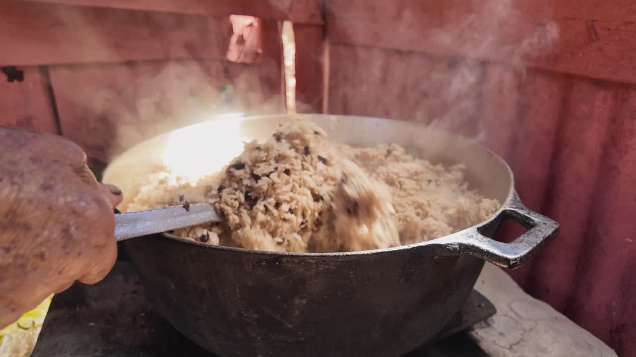 A serene countryside setting where a humble wood stove simmers traditional Dominican moro. The steam rises as the metal spoon stirs, showcasing authentic rural cooking techniques and culture