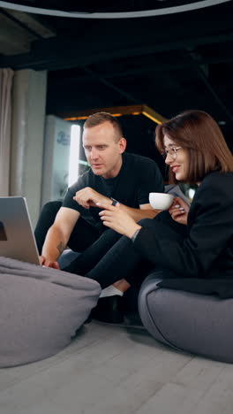 friendly colleagues' communication in the office. man and woman sit in bean bag chairs looking at laptop and discussing. vertical video.