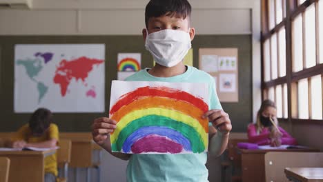 Junge-Trägt-Gesichtsmaske-Und-Hält-Ein-Regenbogengemälde-Im-Unterricht-In-Der-Schule