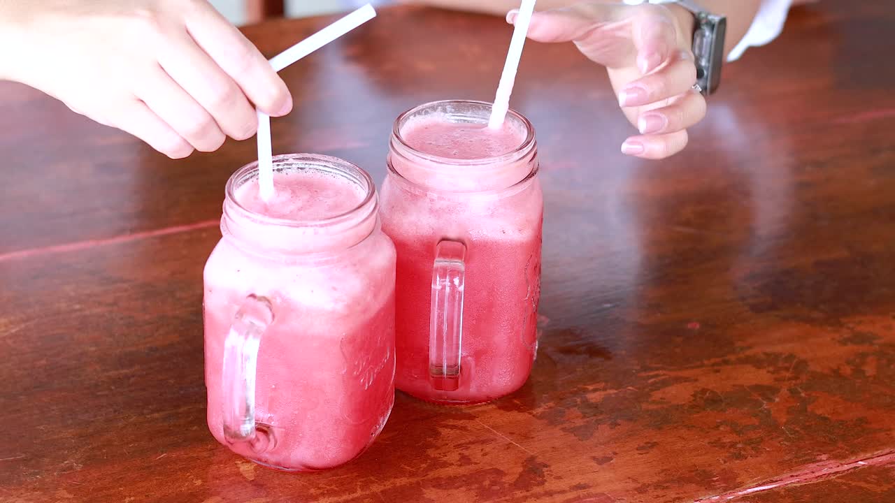 Hands reach for vibrant watermelon juice in mason jars on a wooden table, capturing a refreshing moment in bright, natural lighting